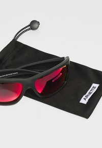 Lunettes de soleil noires avec des verres teintés en rouge, fabriquées à partir de plastique recyclé, reposant sur une pochette en tissu noir arborant un logo "ARNETTE" en blanc.