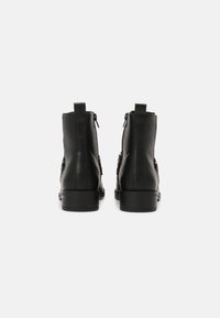 Un par de botas de cuero negro hasta el tobillo mostradas desde atrás, con cremalleras laterales y tacones bajos apilados sobre un fondo blanco liso.