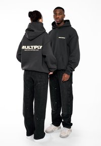 Los sudaderas con capucha negras presentan un gran logo "MULTIPLY" en la parte posterior. Combinadas con pantalones de mezclilla negros, el conjunto resalta el estilo moderno del streetwear.