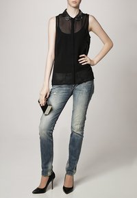 Blusa nera senza maniche con pannelli trasparenti e colletto borchiato, abbinata a jeans skinny blu sbiaditi e tacchi alti neri. Tiene una piccola pochette nera.