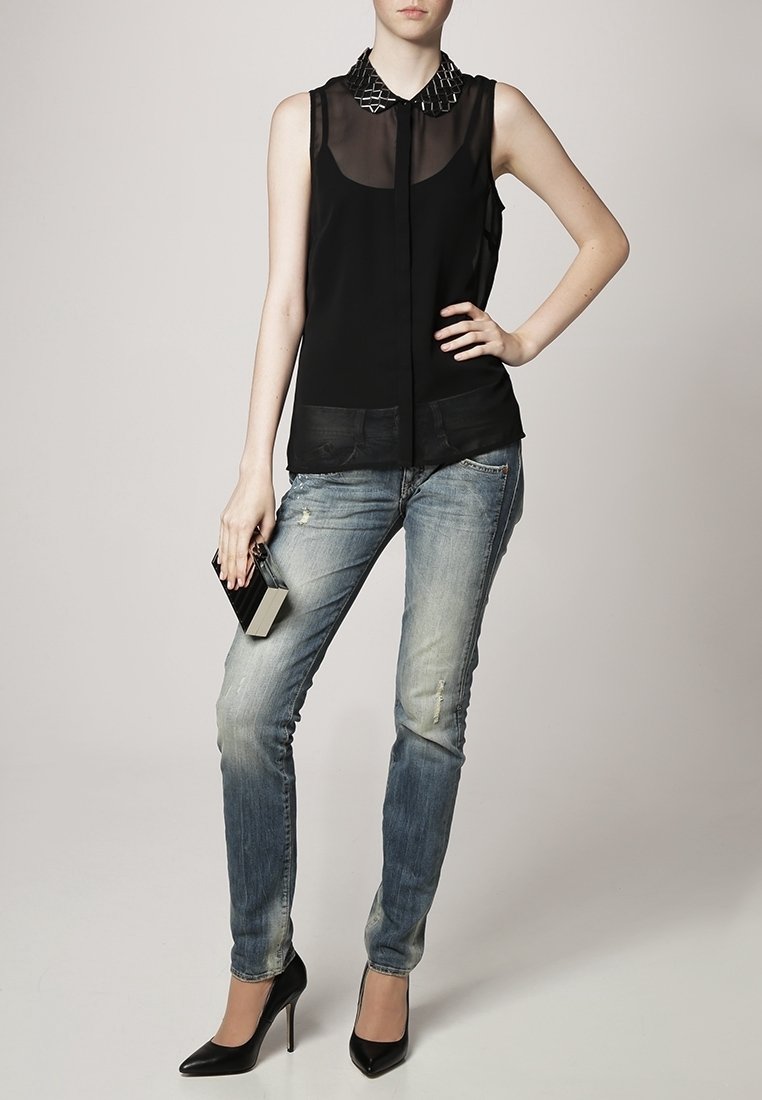 Blusa nera senza maniche con pannelli trasparenti e colletto borchiato, abbinata a jeans skinny blu sbiaditi e tacchi alti neri. Tiene una piccola pochette nera.