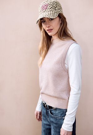 Donna che indossa un berretto con stampa leopardata, un maglione senza maniche rosa pallido sopra una camicia bianca a maniche lunghe e jeans blu, in piedi davanti a uno sfondo beige.