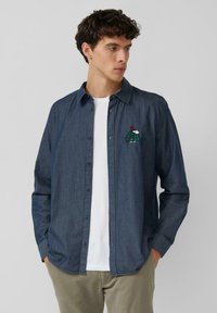 Granatowa jeansowa koszula na guziki z długimi rękawami, z zielono-białym haftowanym wzorem żółwia na lewej piersi.