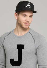 Felpa grigia con una lettera "J" nera sul davanti, con dettagli di cucitura nera. Cappellino nero con un logo bianco "A".