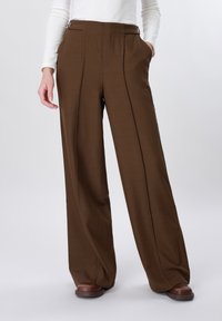 Femme portant un pantalon marron à taille haute et jambes larges avec des plis sur le devant, associé à un haut blanc à manches longues et des chaussures marron.