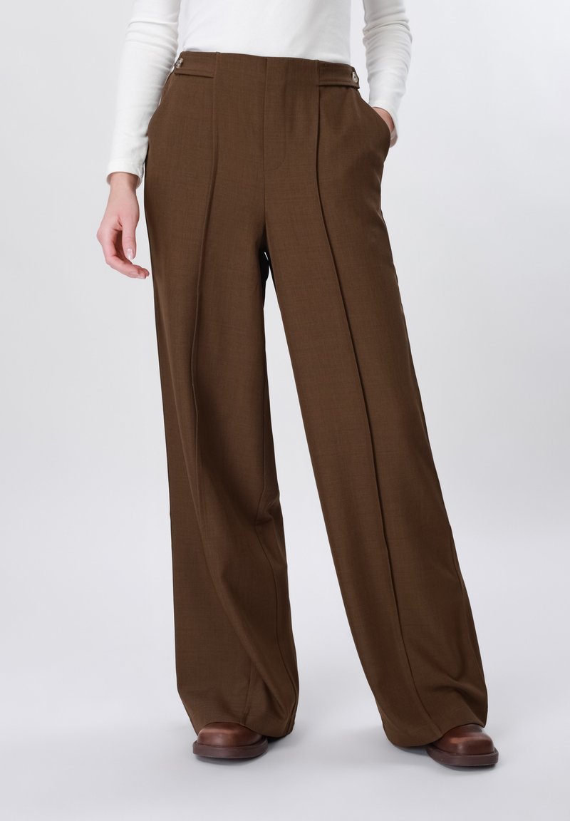 Femme portant un pantalon marron à taille haute et jambes larges avec des plis sur le devant, associé à un haut blanc à manches longues et des chaussures marron.