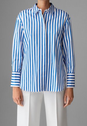 Personne portant une chemise à manches longues rayée verticale bleu et blanc avec de larges poignets et un pantalon blanc, debout devant un fond gris.