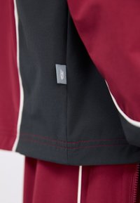 Bordeaux- og marineblå jakke med hvid piping. Stoffet ser glat ud med en subtil tekstur. Har et lille gråt mærke med et emblem.
