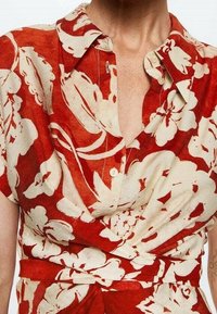 Camicetta avvolgente rossa con fiori, dotata di colletto con bottoni, maniche corte e un tessuto texturizzato con grandi motivi floreali di colore crema.