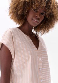 Beżowa bluzka w paski z krótkim rękawem, dekoltem w serek i białymi guzikami. Gładka tekstura z pionowymi białymi paskami na miękkim materiale.