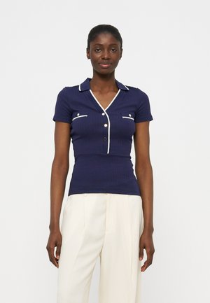 Γυναίκα με εφαρμοστή μπλε-σκούρα κοντομάνικη μπλούζα με λευκή επένδυση και κουμπιά, συνδυασμένη με λευκό παντελόνι ψηλόμεσο και φαρδύ.