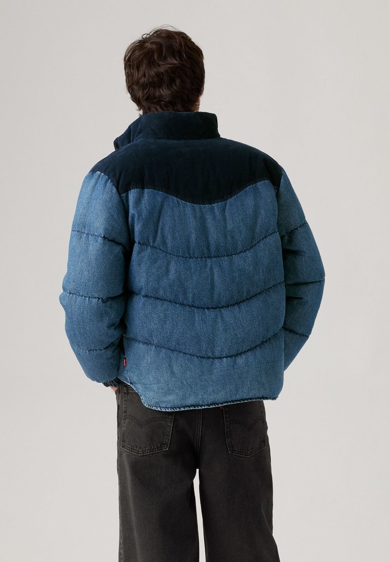 Polstret jakke i varierende nuancer af denimblå med en mørk corduroy-krave, der har et markant quiltet mønster og en afslappet pasform.