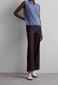 Top de punto texturizado azul sin mangas con cuello alto, combinado con pantalones vaqueros oscuros de pierna ancha y botas de tobillo en color blanco roto. Fondo neutro.