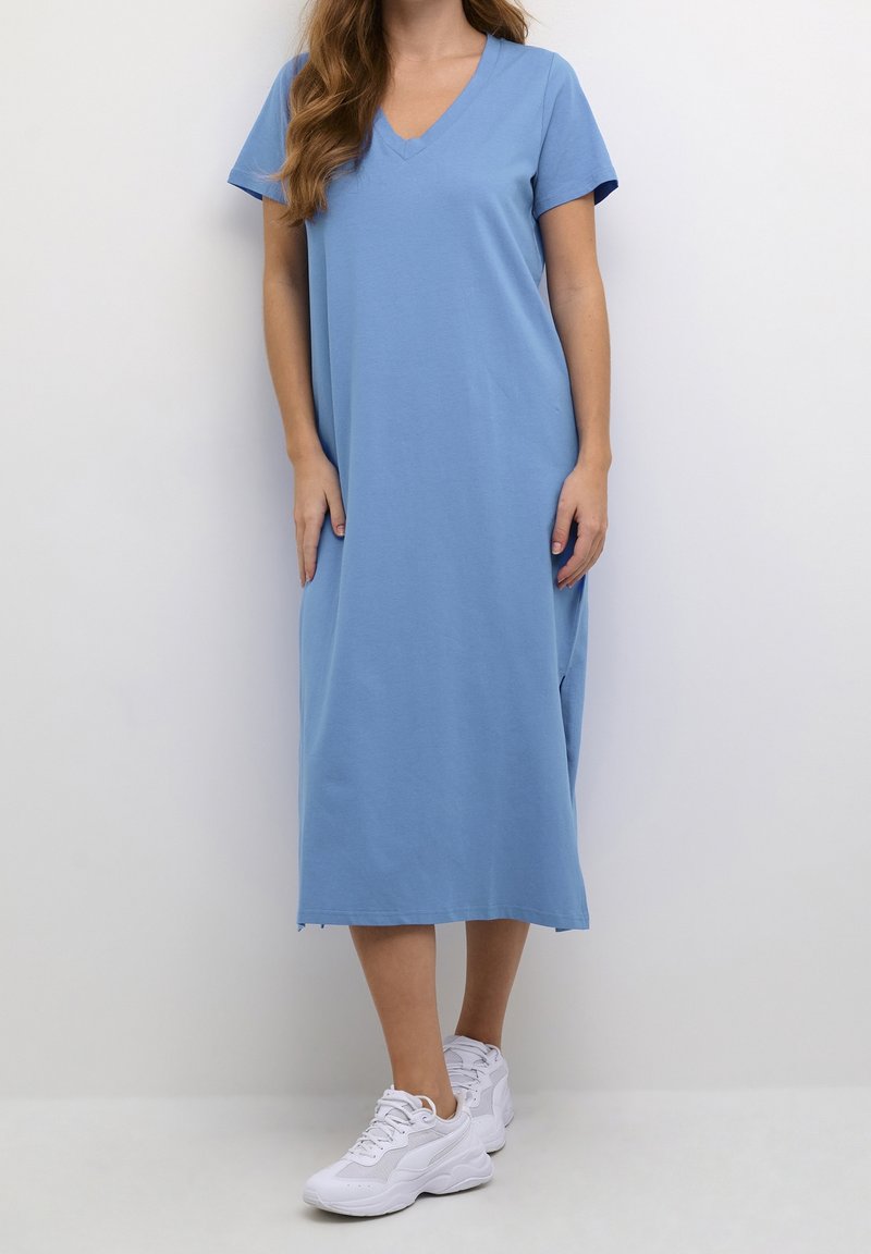 Robe en mélange de coton bleu avec manches courtes, encolure en V et fentes latérales ; longueur midi, associée à des chaussures de sport blanches.