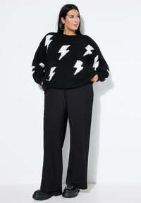 Frau mit langen dunklen Haaren, die einen schwarzen Pullover mit weißen Blitzen, schwarze weit geschnittene Hosen und schwarze klobige Plateau-Schuhe trägt.