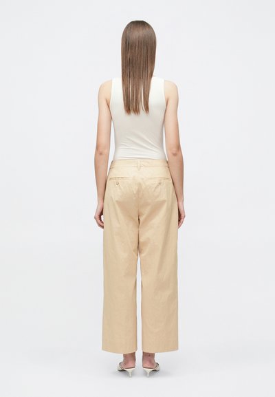 Γυναίκα με ίσια καστανά μαλλιά φοράει αμάνικη λευκή μπλούζα, φαρδιά μπεζ παντελόνια και λευκά σανδάλια με τακούνι, στέκεται γυρισμένη προς τα πίσω σε λευκό φόντο.