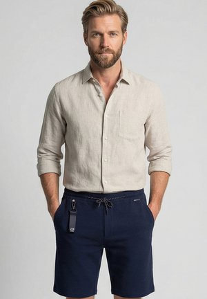 Man met baard, gekleed in een beige shirt met lange mouwen en marineblauwe shorts met trekkoord en een sleutelhanger eraan vastgeklikt, staand met handen in de zakken.
