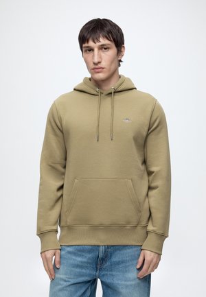 Homme portant un sweat à capuche marron clair avec poche et un jean bleu, debout devant un fond blanc uni.