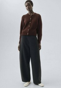 Cardigan en tricot marron avec boutons, associé à un pantalon large gris foncé. Texture lisse, design minimaliste, complété par des chaussures de couleur claire.