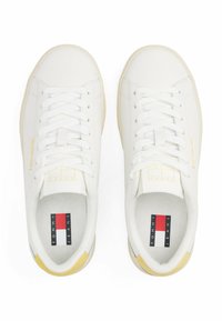 Baskets blanches avec une tige en cuir lisse, des lacets plats, des accents jaunes au niveau du talon, et une semelle intérieure texturée comportant le logo Tommy Jeans.
