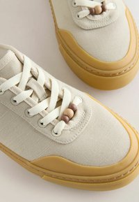 Baskets en toile beige avec des lacets blancs et trois perles en bois décoratives sur chaque chaussure, avec des semelles en caoutchouc couleur fauve et des bouts renforcés.