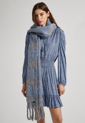 Hnedosivý a modrý prechodový šál s mäkkou, nadýchanou textúrou a strapcovými okrajmi, nosený cez kvetinové modré šaty so smockovaným pásom.