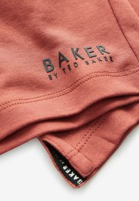 Rustonsävyinen kangas, jossa on pehmeä rakenne. Helman lähellä on musta "BAKER by TED BAKER" -logo ja reunassa musta etiketti.
