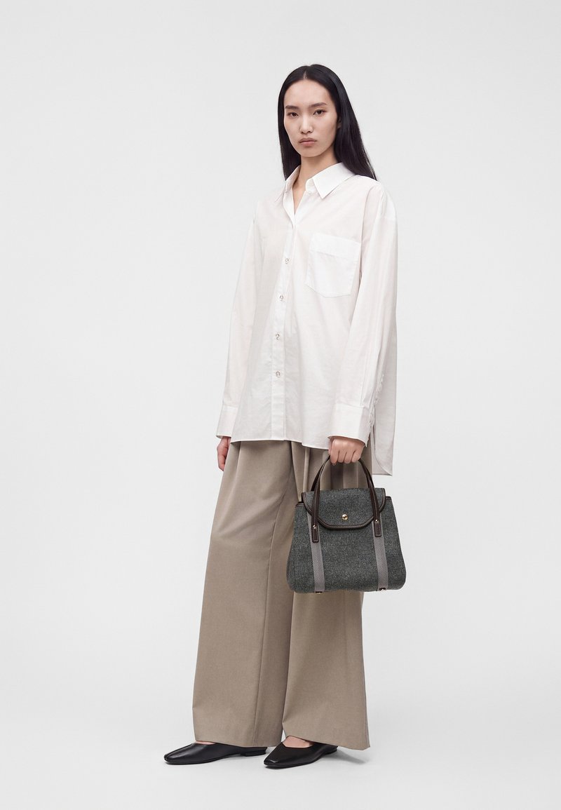 Femme aux longs cheveux noirs portant une chemise blanche ample à boutons et un pantalon large beige, tenant un sac à main gris et portant des ballerines noires.