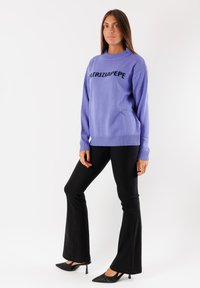 Maglione lavanda con la scritta "PATRIZIA PEPE" in lettere a maglia nera, collo a girocolla a coste e maniche lunghe, abbinato a pantaloni neri a campana e tacchi.