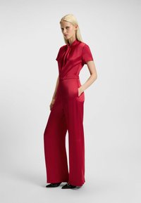 Completo in satin rosso con maniche corte, vita aderente, pantaloni a gamba larga e tasche laterali. Tessuto liscio e dettaglio della cravatta centrale al collo.