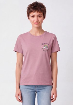 Jeune femme aux cheveux courts et bruns portant un t-shirt mauve avec un petit arc-en-ciel et le texte "Happiness is homemade" sur la poitrine, associé à un jean bleu clair.