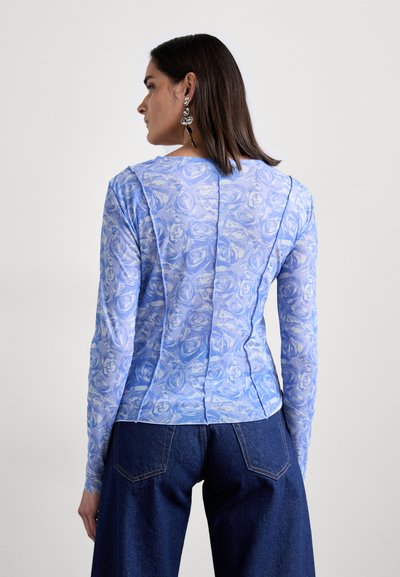 Μακρυμάνικο μπλουζάκι με γαλάζιο λουλουδάτο σχέδιο, φτιαγμένο από διάφανο ύφασμα, με στρογγυλή λαιμόκοψη και λεπτομέρειες ραφών. Φορεμένο με τζιν παντελόνι.