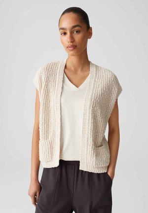 Beige gebreide vest met korte mouwen, open voorkant en twee voorzakken; gedragen over een witte v-hals shirt, gestructureerde stof, losse pasvorm.