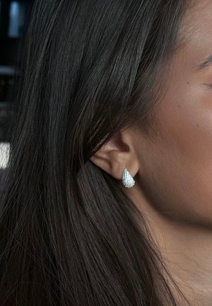 Tårformade örhängen i silverfärgad metall med glimrande, klara rhinestones. Bakgrunden visar mörka, suddiga toner.
