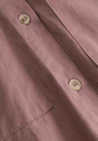 Rožā tekstila tuvplāns ar gludu, matētu virsmu, pogas detaļu un redzamām šuvēm apkārtējiem malām.