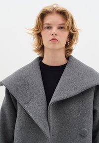 Grau übergroßer Wollmantel mit breitem Revers, zwei Knöpfen und einer weichen Textur, kombiniert mit einem schwarzen Rundhals-Pullover darunter.