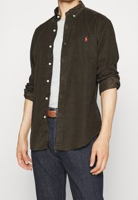 Mann trägt ein unbuttoniertes dunkelbraunes Cordhemd über einem hellgrauen T-Shirt, blaue Jeans und einen braunen Ledergürtel, die Hände entspannt an den Seiten.