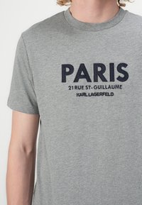 Pilkas medvilnės marškinėliai su jūrų mėlynu siuviniu „PARIS 21 RUE ST-GUILLAUME“ virš „KARL LAGERFELD“, apvalia kaklo išpjova, trumpomis rankovėmis.