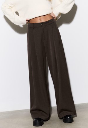 Personne portant un pantalon large noir, des chaussures plateformes noires et un pull court blanc, mains sur les hanches, sur un fond uni.