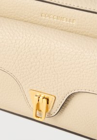 Μπεζ δερμάτινη τσάντα χειρός με υφή στην επιφάνεια, κούμπωμα σε χρυσαφί απόχρωση και λογότυπο σε χρυσό χρώμα. Οι άκρες είναι ραμμένες με κλωστή σε αντίθετο χρώμα.