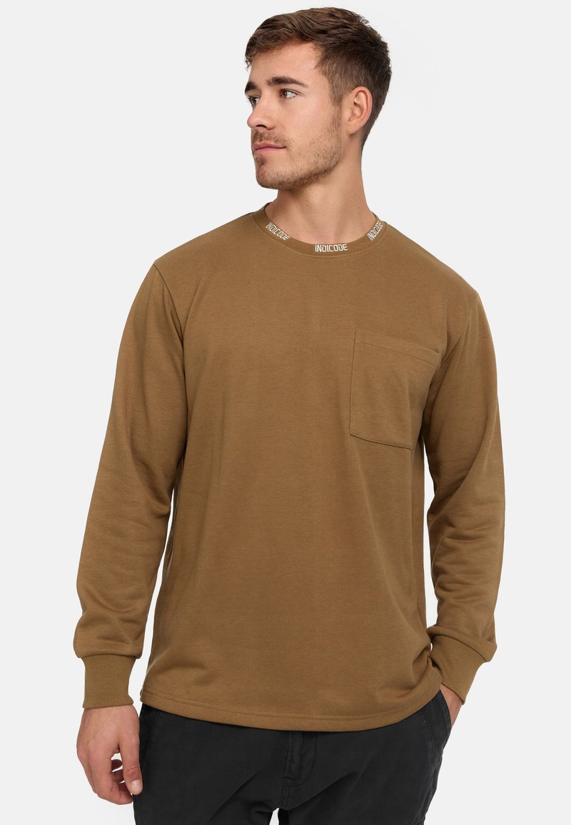 Brun långärmad t-shirt med rund halsringning och en kontrasterande vit varumärkesetikett, en bröstficka och ribbade manschetter.