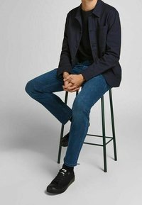 Navy button-up shirt, black t-shirt, blue jeans, and black sneakers worn by a seated person on a green metal bar stool.