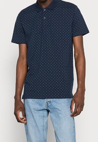 Muž v navy modrém polo tričku s krátkým rukávem a bílým diamantovým vzorem, v světlých modrých džínech, stojící na jednobarevném pozadí.