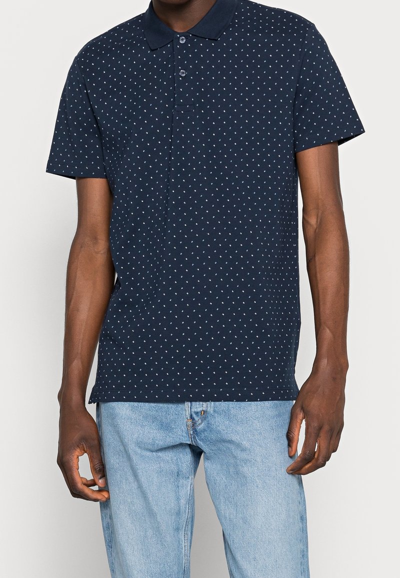 Muž v navy modrém polo tričku s krátkým rukávem a bílým diamantovým vzorem, v světlých modrých džínech, stojící na jednobarevném pozadí.