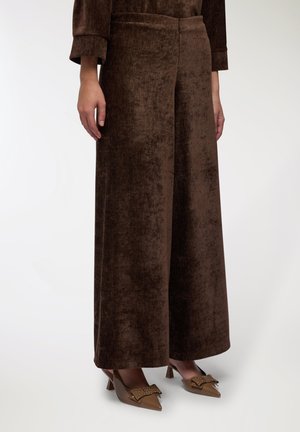 Persona che indossa pantaloni larghi in velluto marroni e tacchi a punta marroni con dettagli in forma di fiocco gioiello, in piedi contro uno sfondo semplice.