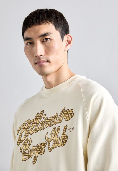 Young man with short dark hair wearing a cream sweatshirt printed with "Billionaire Boys Club" in brown checkered script.