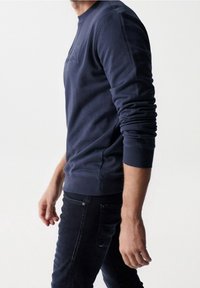 Uomo che indossa una camicia a maniche lunghe blu navy e jeans scuri, mostrato dalla spalla a metà coscia, su uno sfondo chiaro e semplice.