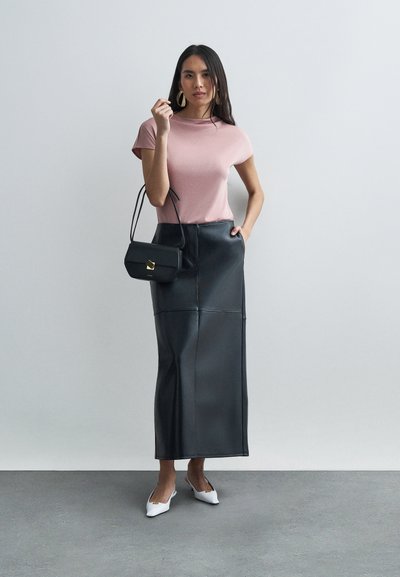 Žena s růžovým tričkem s krátkým rukávem, černou koženou midi sukní, bílými špičatými podpatky, velkými zlatými náušnicemi a držící černou kabelku.