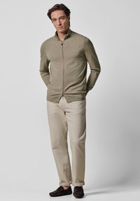 Sweter na zamek w jasnym oliwkowym odcieniu zieleni z długimi rękawami, zestawiony z beżowymi spodniami i ciemnymi butami wsuwanymi. Gładka faktura, casualowy krój.