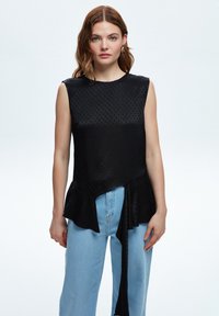Top senza maniche nero con pois a rilievo, orlo a peplum e dettaglio con fiocco laterale, abbinato a jeans a gamba larga di colore blu chiaro.